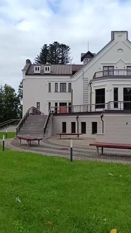 This is a vertical, exterior shot of a large, white, two-story building with a steeply pitched roof and a smaller tower-like structure on top. The building has a balcony with railings on the second story, and a wide set of stairs leading up to the main entrance from a paved courtyard. Benches are placed throughout the courtyard and on the grassy area in the foreground. Trees are visible behind the building and on the right side of the frame. The sky is overcast.