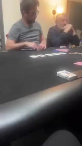 Two men sit at a poker table, playing cards. One man wears glasses and a grey t-shirt, and is in the process of pondering his next move. The other man sits back in his chair, arms crossed and looking at the table. On the table, there are cards, poker chips, and a pack of cigarettes. The table is covered in black felt, and the background is a simple room.