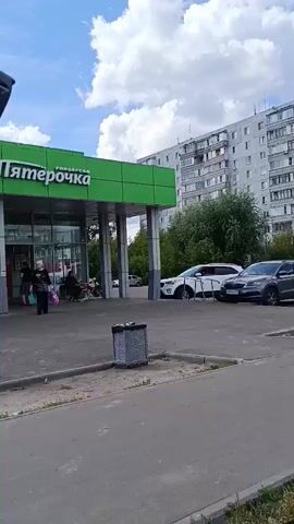 The video opens with a view through an open car door of a Peryochka grocery store. People are seen entering and exiting the store, and a motorcycle is parked near the entrance. The camera then pans to the right, showing a row of parked cars and a sidewalk. A man walks along the sidewalk carrying a shopping bag. The camera continues to pan right, revealing more parked cars and a billboard advertising "Femto-LASIK" eye surgery. Pedestrians are visible near the billboard. The video ends with a view of more parked cars and a street sign. The weather appears to be sunny with some clouds.