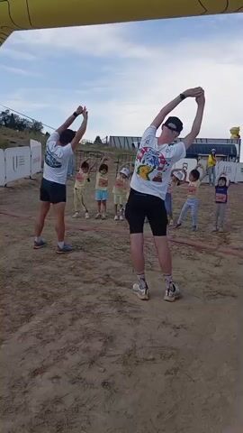 Two men and several children are stretching under a yellow archway. The men are in the foreground, facing away from the camera. They are wearing t-shirts and shorts. The children are in the background, also stretching. The environment appears to be an outdoor event, possibly a race or a sporting event, with a dirt ground and some fencing in the background. The sky is cloudy.