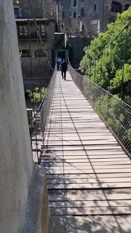 The video shows a person walking across a wooden suspension bridge. The bridge has metal railings and appears to be old. In the background, there are stone buildings and trees. The sky is blue and the sun is shining. Another person is walking in front of the first person. The camera is positioned on the bridge, looking forward towards the buildings. The first person is walking away from the camera.