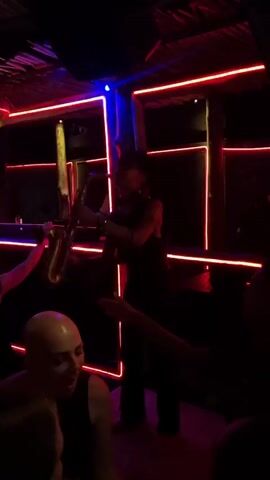 The video depicts a vibrant nightlife scene inside what appears to be a bar or club. A saxophonist in dark clothing stands on a platform, illuminated by red neon lights, as he performs for the crowd. The room is dimly lit with disco balls hanging from the ceiling and a "no worries" sign visible in the background. People can be seen dancing and raising their hands in the air, enjoying the music. The scene is lively, with the focus on the music and the energetic atmosphere of the venue.