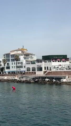 The video pans from right to left, showing a waterfront scene. On the left, a white building with multiple stories and balconies overlooks a rocky shoreline and the water. A red buoy is visible in the water. In the center, a tan stone tower with arched windows stands near a white building with a green, vegetated roof and outdoor seating. Flags are flying from poles near the tower. Further to the right, a walkway juts out into the water, lined with rocks and palm trees. Several large white yachts are docked behind the walkway, with people visible on the deck of one. In the distance, a mountain range forms the backdrop. A small boat with two people in it is moving across the water in the foreground. The sky is clear and blue.
