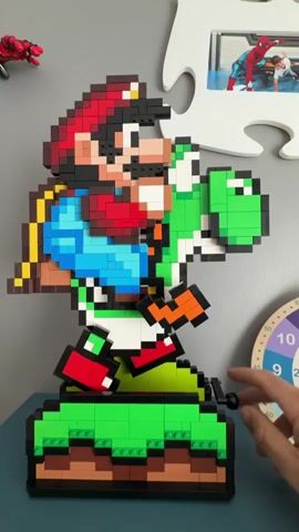 A person's hand is attaching black lego bricks to a large, pixelated lego sculpture of Mario riding Yoshi. The sculpture is mostly red, green, blue, and black, and is standing on a base made of stacked brown and green legos that resemble grassy hills. In the background, there is a gray wall, a white puzzle piece frame, and other objects like a Spiderman figurine and a clock. The hand moves, attaching the bricks to the base of the sculpture. The person then picks up the entire sculpture, turning it slightly to showcase its detail.
