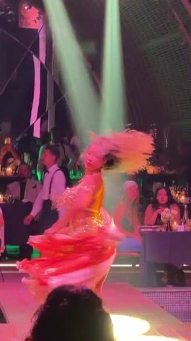 A woman is belly dancing in what appears to be a restaurant or club. She is wearing a gold and orange outfit and has long blonde hair. The room is decorated with modern, geometric structures. Tables with people are visible in the background, many watching the dancer.  Green and pink spotlights illuminate the dancer as she moves around the stage.