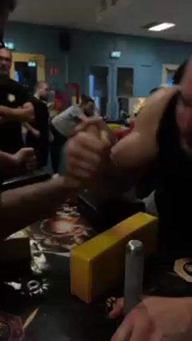 The video depicts two men arm wrestling in a room. One man wears a black tank top and has a tattoo on his left bicep. The other man wears a black t-shirt. They grip hands tightly on a table designed for arm wrestling with yellow pads. Several other people stand around watching, including a man with glasses and a black t-shirt who stands directly in front of the camera. The room has blue walls and multiple doors and windows. A red fire extinguisher is visible in the background.