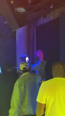 The video shows four men in a nightclub or event space with purple and yellow lighting. One man is playing a set of timbales drums, while another stands beside him. The other two men are standing in front, facing the drums, and appear to be watching or dancing. The man playing the timbales has a pink afro-style wig. The men are casually dressed and appear to be having a good time. The environment looks like an indoor party or concert with sound equipment visible in the background.