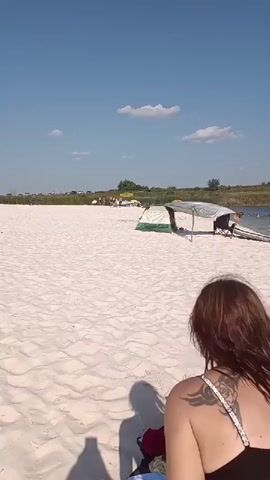 The video opens with a view of a sandy beach under a bright blue sky with scattered clouds. In the foreground, the back of a person wearing a dark, patterned tattoo on their shoulder is visible. The camera then pans across the beach, revealing a tent set up with a tarp overhead. Several people are visible further down the beach near the water, some appearing to be swimming or relaxing. Umbrellas and beach gear are scattered around. Cars are parked in the background, suggesting a parking area. The camera then focuses on a person sitting on the sand, wrapped in a towel, with a phone placed on a blue towel beside them. There are several items of clothing on the sand near this person. The video ends with a close-up of the person sitting on the sand.