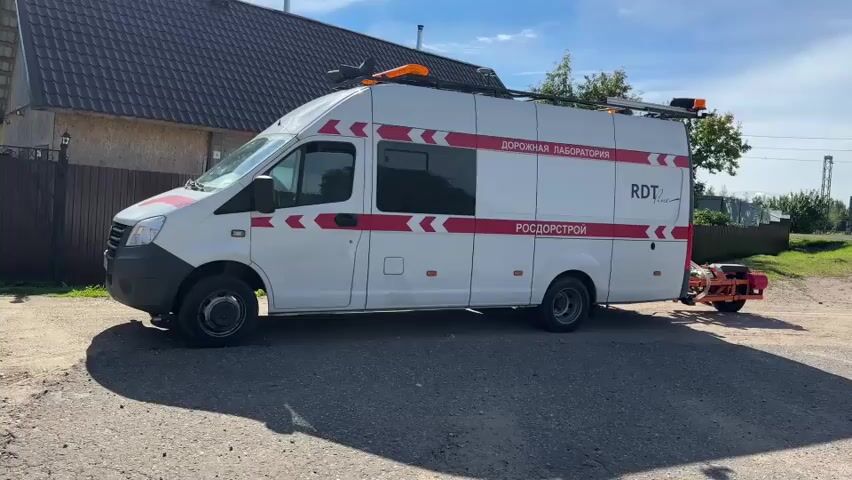 A white van with red stripes and the words "РОСДОРСТРОЙ" and "ДОРОЖНАЯ ЛАБОРАТОРИЯ" in Russian is parked on a dirt road next to a paved road. The van has equipment mounted on its roof and a trailer attached to its rear. The trailer has orange and black equipment on it. The van is casting a shadow on the ground. In the background, there is a house with a dark roof, a fence, trees, and a blue sky with clouds.