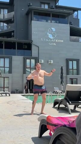 A man in swim trunks dances by a pool in front of a building with the words STILLATERM SPA. He moves his arms and torso, with his head tilted slightly. The pool is surrounded by lounge chairs and a person can be seen in the background near a doorway.