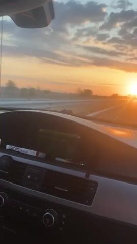 The video shows the interior of a car as it drives down a highway during sunset. The sun is low on the horizon, casting a warm, orange glow across the sky and the road. The car's dashboard is visible in the foreground, with a navigation screen displaying a message in German. The road ahead appears to be empty, with a few trees visible in the distance. The sky is filled with dramatic clouds, which are illuminated by the setting sun. The overall mood of the video is peaceful and serene, with a sense of motion and journey.