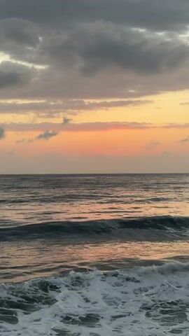 This is a close-up, vertical shot of a turbulent ocean under a cloudy sky at sunset. The sky displays a gradient of colors, from a pale orange near the horizon to gray clouds higher up. Waves are crashing against the shore in the foreground, with white foam created by their impact. The water is dark and choppy, reflecting the colors of the sunset.