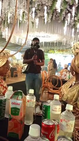 A man holding a camera on a gimbal stands in the foreground, facing a mirror that reflects him and a woman wearing sunglasses and a headscarf. The background shows a decorated event space with tables and chairs, possibly a wedding reception. On the table in front of the mirror are several bottles of water, cans of soda, and a carton of orange juice. The man is filming the scene, with the mirror acting as a visual intermediary. The woman is seated and looking towards the mirror.