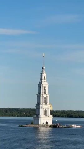 A tall, white bell tower stands in the middle of a body of water. The tower has multiple levels with arches and windows, and a cross at its peak. A small island or platform is attached to the base of the tower, with a dock and several people standing on it. A small boat is docked at the platform. In the background, a line of trees forms the shoreline, and a faint golden dome is visible behind the trees. The sky is clear and blue with a few wispy clouds.
