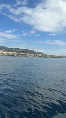 The video shows a coastal city from the perspective of a boat out on the water. The water is blue and rippled, with a city skyline visible on the horizon against a backdrop of mountains. The sky is blue with scattered white clouds. The camera moves slightly, revealing parts of the boat's structure, including a metal railing and the edge of a sunshade.