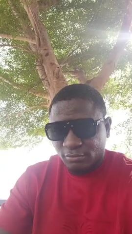 A man with dark skin and sunglasses is looking at the camera. He is wearing a red shirt. Behind him is a tree with green leaves and sunlight filtering through. He holds up two fingers in a peace sign.