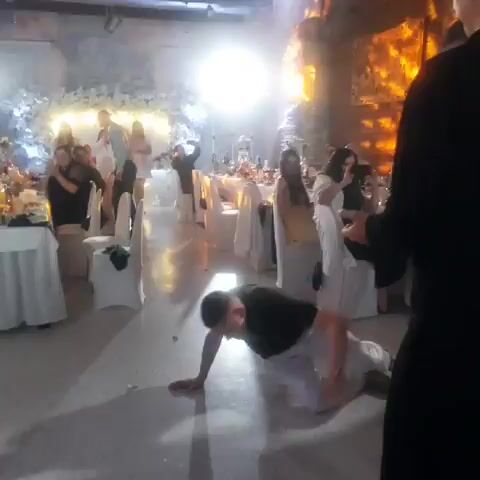 The video shows a man breakdancing at what appears to be a wedding reception. He is wearing a black t-shirt and white pants and is in the middle of a move, with his legs in the air and his hands on the floor. The room is decorated with white tablecloths and chairs, and there are people seated at tables in the background. There is a bright spotlight shining on the dance floor, and the room is dimly lit otherwise. The man appears to be alone on the dance floor.