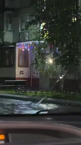 This is a nighttime view from inside a vehicle looking out at a building's entrance. The building is a multi-story structure, possibly an apartment building, with windows visible on upper floors. The entrance area is decorated with colorful string lights. A bench and possibly a bicycle are visible near the entrance. A street light illuminates the area. The foreground shows a portion of the vehicle's interior, including part of the dashboard with an illuminated display. There are also water droplets on what appears to be a windshield. The overall atmosphere is dark and somewhat damp. No people are visible in the frame.