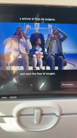 The video displays an airplane safety demonstration on a screen. Three people, two adults and one child, are shown on the screen wearing oxygen masks. The adult on the left is a man with dark skin, wearing a light-colored jacket. The adult on the right is a woman with blonde hair, wearing a striped shirt and a jacket. The child is sitting between them, holding a teddy bear. The text on the screen is in Spanish and English, providing instructions on how to use the oxygen masks. The English text reads: "Place the mask over your nose and mouth, slip the elastic band over your head and tighten the straps." The video appears to be paused, as indicated by the "PA in progress" text at the bottom right of the screen. The foreground of the image shows a part of the airplane's interior, including a white armrest.