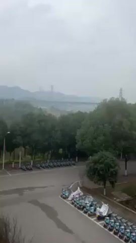 The video shows an exterior shot of a street scene on a cloudy day. The foreground features a paved area with parked motorcycles, some covered with tarps. Beyond the paved area is a row of trees and a road. In the background, a large electrical substation and a bridge structure are visible, shrouded in mist. Mountains are faintly discernible in the far distance. There is no apparent human activity.