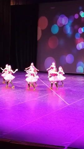 Six young girls in white and pink tutus with roller skates on their feet are performing a dance routine on a stage. The stage is lit with bright purple lights. Behind the dancers, a screen displays a colorful bokeh pattern of circles in various sizes and colors, including pink, red, blue, and purple. The background behind the screen is dark. The girls are spread out across the stage, some facing the audience, others facing each other. They appear to be in motion, with some of them twirling. The overall atmosphere is festive and energetic.