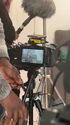 The video shows a behind-the-scenes view of a video shoot. The foreground is dominated by two professional video cameras on tripods, both capturing the same scene which is visible on their monitors: a woman sitting on a sofa in a living room setting, appearing to be the subject of an interview or recording. A person, possibly a camera operator or assistant, is visible on the left of the frame, adjusting a cable or connection on one of the cameras. Another person holding a boom microphone is standing to the left, positioned to capture sound. The room setting includes a coffee table with plants, a gray sofa, and decorative elements, suggesting a comfortable and well-lit environment for the interview. The monitors on the cameras display technical information, including timecodes and recording parameters. The presence of multiple cameras and the boom microphone indicate a professional video production setup.