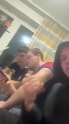 Three young men are in a room, seated on the floor. The man closest to the camera wears a black hoodie and sticks his tongue out. The middle man wears glasses and a red and blue shirt. The man farthest from the camera looks at his phone. A yellow and white curtain hangs in the background. There is also a desk with objects on it visible in the background.
