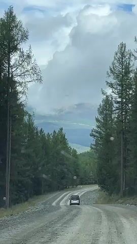 A vehicle drives down a gravel road through a forest. The road curves to the right and leads into a valley. The sky is cloudy with some patches of blue. The trees are tall and green, and the mountains in the distance are covered in mist. The video is taken from inside another vehicle, and there are several water spots visible on the windshield. The time stamp on the video ranges from 00:00 to 00:05.