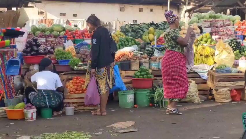 The video shows a bustling outdoor market scene. Various vendors display a wide array of fruits and vegetables, including watermelons, tomatoes, eggplants, avocados, bananas, and leafy greens. Customers, mostly women, are browsing and shopping, dressed in a variety of clothing, many wearing traditional African prints. A motorcycle passes through the foreground. The market stalls are constructed from wooden crates and tables. Some customers carry bags, presumably containing their purchases. In the background are buildings, possibly shops or residences, adding to the crowded and vibrant atmosphere of the market.
