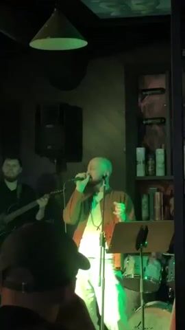 A group of musicians are performing on a stage in a dimly lit room. A bald man in the center is singing into a microphone while wearing a light brown jacket over a white t-shirt and light-colored pants. To his left, a man with a beard plays an electric guitar. To the right of the singer, a drum set is visible, and a woman with dark hair walks by in the foreground. The stage is lit with a combination of blue, red, and white lights. Shelves with bottles and books are visible in the background.