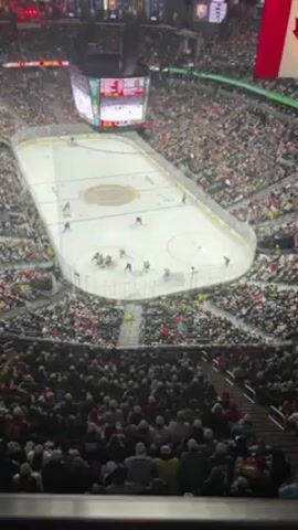 The video shows a hockey game in progress in an arena filled with spectators. The game is being played on an ice rink marked with lines and circles. There are multiple players on the ice, wearing either black or white uniforms with helmets. They are skating around the rink, holding hockey sticks, and interacting with each other. A large scoreboard is visible overhead, displaying the game's statistics. The arena is packed with people in the stands, all watching the game. Some spectators are wearing white hats. The overall setting is a high-energy sporting event.