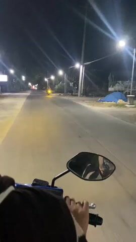 The video shows a first-person perspective of someone riding a motorcycle at night. The road is illuminated by streetlights and the headlights of other vehicles in the distance. A yellow motorcycle is visible approaching in the opposite direction in the early frames. The rider's hands are on the handlebars, and a side mirror reflects the rider and the surroundings. The environment appears to be a rural or suburban area with trees and buildings lining the road. The overall mood is calm and quiet.