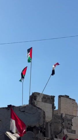 The video shows a partially destroyed building with several flags flying above it. The flags appear to be national flags, including the Palestinian flag and the Egyptian flag. The building's facade is heavily damaged, with exposed rebar and rubble. The sky is clear and blue. In the foreground, there is a sandy area, possibly a construction site, with a fence made of white sheeting in front of the building. Several other flags are visible in the distance, strung along a line. The overall scene suggests a damaged urban environment, possibly in an area affected by conflict.