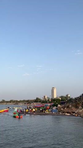 A vertical, medium-shot video shows a bustling beach scene from the perspective of the water. In the foreground, the dark blue water is slightly choppy, with a few small boats visible. One boat, painted green and red, is in motion near the center, with people inside. Another boat, bright yellow, is further to the left, also with people. The middle ground is the beach itself, which is crowded with people. There are numerous colorful tents and stalls set up, suggesting a market or vendor area. People are walking around, some appearing to be shopping or interacting with vendors. In the background, a tall, light-colored building stands out against the pale blue sky. Several other smaller buildings and trees are also visible, hinting at a developed area behind the beach. The sky is clear, with a few wispy clouds. The overall impression is of a lively and active recreational area.