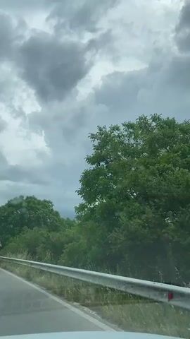 The video, captured from a moving vehicle, displays a rural road scene under a heavily overcast sky. The road's asphalt surface is visible on the left, bordered by a silver guardrail. Beyond the guardrail, lush green trees and overgrown grass line the roadside. Dark, heavy clouds dominate the sky, suggesting an impending rain or gloomy weather.

At approximately the 0:02 mark, a blue road sign comes into view on the left. It features white text in Cyrillic, indicating directions and distances. The upper line reads "Асвовница" (Asvovnitsa) with an arrow pointing left and the number "6" (likely kilometers). The lower line reads "Добрашница" (Dobrashnitsa) with a straight arrow and the number "2" (likely kilometers). As the vehicle continues, the sign passes, and the view returns to the winding road, the guardrail, and the dense foliage, with distant mountains faintly visible beneath the heavy cloud cover. The overall impression is one of travel through a scenic, yet somber, natural landscape.