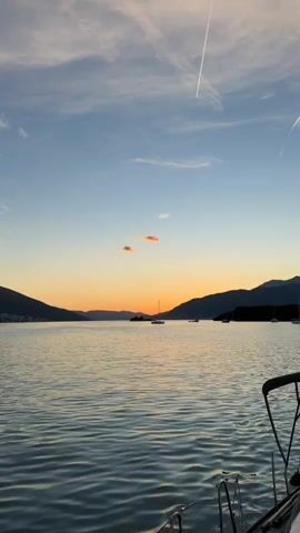 The video shows a serene sunset over a body of water. The sky is a gradient of blue and orange, with a few wispy clouds and a contrail from an airplane. Silhouetted mountains frame the horizon, and several sailboats are anchored in the distance. The foreground is occupied by the railing and deck of a boat, implying the viewer is on board and looking out at the tranquil scene. The water is calm, reflecting the colors of the sky. The overall impression is one of peace and natural beauty.