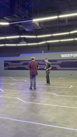 Two individuals are wearing VR headsets in a large, indoor space with a grey floor marked with white lines creating geometric shapes. The person in the foreground wears a maroon t-shirt and blue jeans, holding small objects in their hands and looking downwards. The second person is a few steps behind and slightly to the right, wearing a grey shirt and dark pants. A wall in the background is painted grey, with a futuristic-looking design in black and silver and the word "WARPOINT" written in bold. Overhead lighting consists of strips of fluorescent lights, and the area appears to be part of a VR gaming facility.