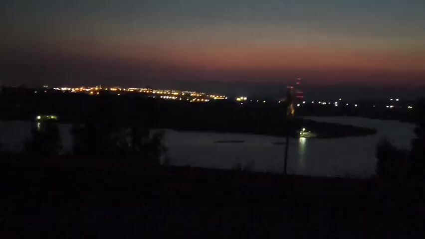 A wide shot captures a twilight landscape featuring a body of water in the foreground and a city skyline in the distance. The sky is a gradient of dark blue and purple, transitioning to orange near the horizon. Numerous lights from the city twinkle across the distant landscape, suggesting a populated area. The water reflects some of these lights, creating subtle ripples of light on its surface. Silhouettes of trees are visible in the extreme foreground, obscuring the lower portion of the frame. The overall scene is dark and atmospheric, with the city lights providing the primary source of illumination.
