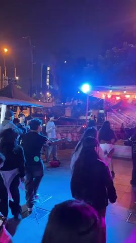 A night time street scene shows people dancing to music in front of a small stage set up outdoors.  A person stands on the stage and sings into a microphone. Behind them is a backdrop with colorful cartoon-style images and text.  Above the stage are strands of lights. The stage area is lit by red, blue and green stage lights.  The dancers are a mix of genders and ethnicities.  They are dressed in casual clothing including hoodies, jeans, and jackets. A street light illuminates a tree to the right of the stage. Cars can be seen driving on the street behind the stage.