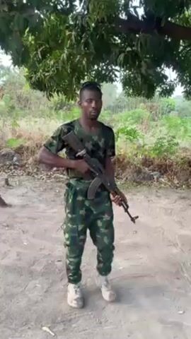 A man dressed in camouflage fatigues is standing beneath a tree holding a rifle. He has dark skin and a close-cropped haircut. He is wearing a short-sleeved camouflage shirt, camouflage pants, and white combat boots. He also has a black shoulder bag. He is holding an AK-47 rifle with his right hand across his chest. He is looking to his left. The scene is outdoors, and the ground is a dirt path. There is lush green vegetation in the background. Some discarded items can be seen on the ground behind the man. There is camouflage clothing draped over a branch of the tree behind him.