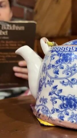 A man is reading a book titled "The Buddhism of Tibet." He has fair skin, blue eyes, and his dark hair is pulled back. He is sitting in front of a brick wall. In the foreground is a blue and white patterned teapot on a wooden table. The teapot is slightly out of focus.