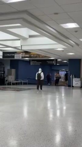 The video shows the interior of an airport. Several people are walking through a wide hallway with a speckled tile floor.  A person wearing a white head covering walks away from the camera, while others pull suitcases past the camera. Airport signage indicates directions to departures, baggage claim, and ground transportation. A sign for the Cleveland Police is also visible. The ceiling has a grid of recessed lighting.