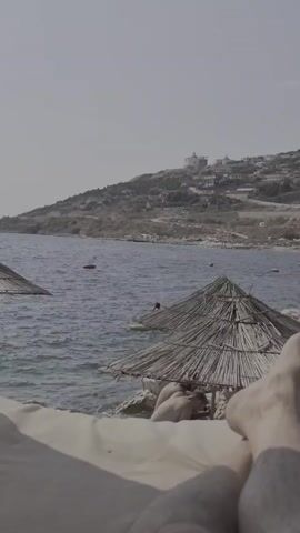 The video opens with a first-person perspective of someone relaxing on a lounge chair, with their feet visible in the foreground. The scene then pans to reveal a picturesque beach with calm blue water and a rocky shoreline. Several thatched-roof umbrellas are scattered across the beach, providing shade. In the distance, a hill overlooks the coastline, dotted with buildings. The water appears relatively clear, and a few people can be seen swimming or lounging on the beach. The sky is a hazy, light blue, suggesting a sunny day. The overall atmosphere is one of relaxation and leisure.