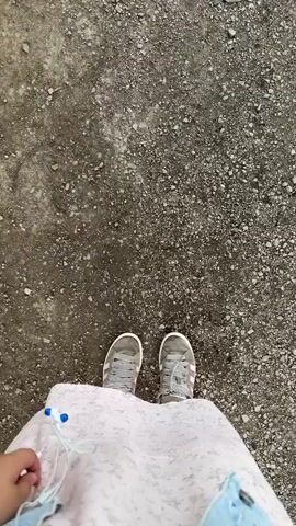 The video shows a first-person perspective of a person walking on a rough, gravelly surface. The person is wearing gray sneakers with white laces and Adidas stripes. They are also wearing a light-colored dress with a floral pattern and a light blue denim jacket. The person is holding white earbuds with blue accents in their left hand. The camera angle is looking down at the person's feet and the ground ahead as they walk. The environment appears to be outdoors, possibly a path or unpaved area. The lighting is natural, suggesting it's daytime. The video is short, showing a few steps being taken.