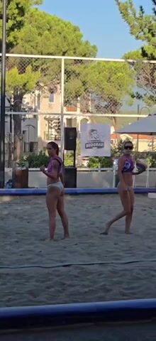 Two women are on a sand volleyball court. The woman on the left is wearing a purple top and white bottoms. The woman on the right is wearing a purple and blue top and white bottoms and is holding a volleyball. She is facing away from the camera. The background shows trees, buildings, and banners with logos. The video appears to be taken during the daytime.