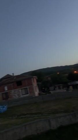 This is a vertical video of a suburban landscape during twilight. In the foreground, there is a low concrete wall and some patchy grass. Beyond that, there are several houses with red-tiled roofs and some with illuminated windows. One house on the left is brick. In the middle ground, there are more houses and some trees. In the background, there are hills and mountains, and a few taller apartment buildings. The sky is a gradient of deep blue at the top transitioning to a pale orange and pink near the horizon. There are streetlights visible, indicating a populated area. The video is shaky and appears to be handheld.