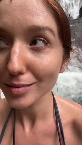 A woman with red hair and freckles stands in front of a waterfall, smiling at the camera. She is wearing a swimsuit top. Other people can be seen behind her, near the waterfall. There is a small bug on her forehead.