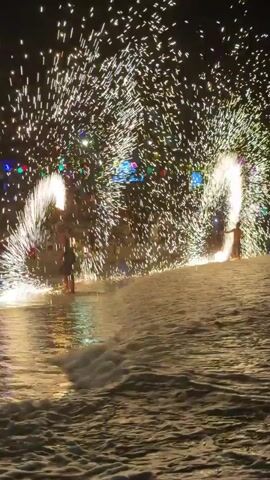 At night, two individuals perform with fire props in shallow water on what appears to be a beach. Each performer holds an object that emits a continuous, fountain-like shower of bright golden sparks, arcing upwards and outwards. The sparks illuminate the performers and reflect vividly on the gently rippling water in the foreground. Distant, multicolored lights are visible in the background.