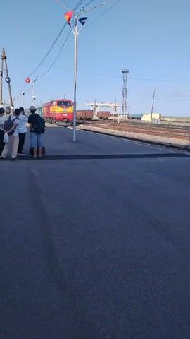 A group of people stand on a paved area next to a train track. A train with a red and yellow locomotive is approaching the people. The train has several blue passenger cars attached to it. The train is traveling on the right side of the frame. The sky is clear and blue. There are power lines and a light pole visible in the image. There is also some industrial equipment in the background on the left side of the frame. The people are facing the train and appear to be waiting for it. The paved area appears to be a platform or a road. The overall scene suggests a train station or a stop along a railway line.