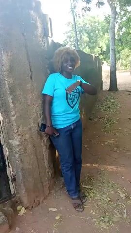 A woman stands near a mud brick wall in a rural setting. She is wearing a blue T-shirt with an emblem on the front that says "THE BEST OF 3," blue jeans, and sandals. She is leaning against the wall, her left hand holding a cell phone and her right hand on her hip. She has a watch on her left wrist. Her hair is light brown and styled in an afro. The ground is dirt with scattered leaves and debris. A few trees are visible in the background. She laughs.
